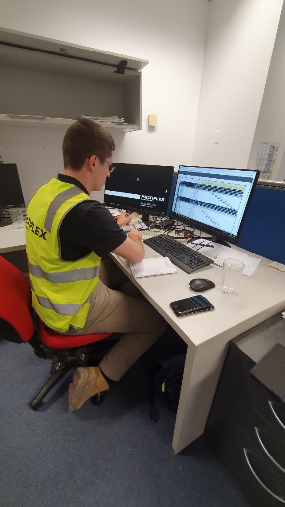Site supervisor at a dual-monitor desk with sticky notes and two phones, reconciling field notes into a spreadsheet.